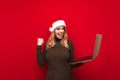 Portrait of a joyful girl with a laptop in her hand, wears a santa claus hat and sweater, rejoices in victory with her hand raised Royalty Free Stock Photo