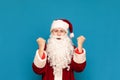 Portrait of joyful guy in Santa suit rejoices with raised hands on blue background, looks into camera and smiles. Young Santa