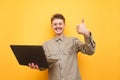 Portrait of joyful nerd in glasses and shirt with laptop in hand on yellow background, looks into camera and shows thumbs up. Royalty Free Stock Photo