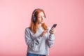 Portrait of a joyful redhead girl who listens to music on headphones with her smartphone in the studio on a pink background. Royalty Free Stock Photo