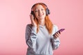 Portrait of a joyful redhead girl who listens to music on headphones with her smartphone in the studio on a pink background. Royalty Free Stock Photo