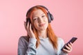 Portrait of a joyful redhead girl who listens to music on headphones with her smartphone in the studio on a pink background. Royalty Free Stock Photo