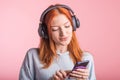 Portrait of a joyful redhead girl who listens to music on headphones with her smartphone in the studio on a pink background. Royalty Free Stock Photo