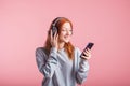 Portrait of a joyful redhead girl who listens to music on headphones with her smartphone in the studio on a pink background. Royalty Free Stock Photo
