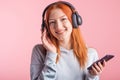 Portrait of a joyful redhead girl who listens to music on headphones with her smartphone in the studio on a pink background. Royalty Free Stock Photo