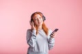 Portrait of a joyful redhead girl who listens to music on headphones with her smartphone in the studio on a pink background. Royalty Free Stock Photo