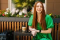Portrait of joyful redhead young woman sitting at table with coffee cup and mobile phone in outdoor cafe terrace in Royalty Free Stock Photo