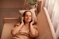 Portrait of joyful relaxed caucasian young woman listening music while resting on couch at home, keeps eyes closed, being calm and Royalty Free Stock Photo