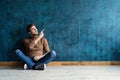 Portrait of a joyful young man sitting on a floor and pointing finger away  over dark blue background. Royalty Free Stock Photo