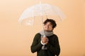 Portrait of joyous african american guy wearing sweater and scarf standing under umbrella, isolated over beige background