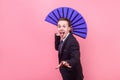 Portrait of joyous carefree young man, magician in elegant suit sticking out tongue and holding big blue fan. on pink