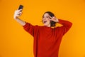 Portrait of joyous woman holding and taking selfie photo on cell phone while standing isolated over yellow background