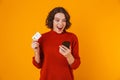 Portrait of joyous woman using cell phone and credit card while standing isolated over yellow background