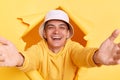 Portrait of kind pleased man wearing casual hoodie and panama looking through torn hole in yellow paper, giving free hugs with Royalty Free Stock Photo