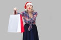 Portrait of kindly beautiful adult woman in red santa cap and checkered shirt standing, holding shopping bags and sending you air Royalty Free Stock Photo
