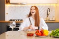 Portrait of attractive redhead young woman with mad eyes holding big knife sitting at table with cutting board and Royalty Free Stock Photo