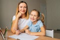 Portrait of laughing elementary daughter doing homework with young mother sitting at home table by window, looking at Royalty Free Stock Photo