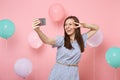 Portrait of laughing young woman in blue dress doing selfie on mobile phone showing victory sign on pink background with Royalty Free Stock Photo