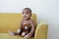 Portrait of Little African newborn baby girl wear diaper smiling and holding brown bear doll while sitting on yellow sofa at home