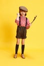 Portrait of little artisitc girl, child in retro shirt and shorts, holding slingshot against yellow studio background