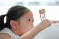 Portrait of little Asian girl looking at hourglass in hand lying on bed at home. Waiting times with sandglass. Close-up shot