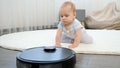 Portrait of little baby boy sitting on carpet at house and looking on working robot vacuum cleaner Royalty Free Stock Photo