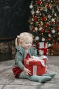 Portrait of little blonde three-year-old girl with big red gift. Christmas tree and garlands background. Xmas mood