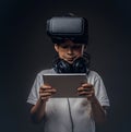 Portrait of a cute little boy dressed in a white t-shirt, with virtual reality glasses and headphones is holding a Royalty Free Stock Photo