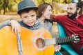 Portrait of little boy in hat playing guitar outdoors. His parents at a background Royalty Free Stock Photo