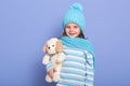 Portrait of little charming girl in knitted winter hat with pom pom, scarf, and striped shirt, holding her favourite soft dog toy Royalty Free Stock Photo