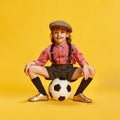 Portrait of little cute girl, child in shirt and shorts posing with football ball against yellow studio background Royalty Free Stock Photo