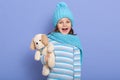 Portrait of little cute girl with dark hair wearing stripped casual shirt, scarf and cap with pom pom, standingagainst blue studio