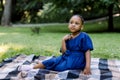 Portrait of little cute happy African girl in blue dress, sitting on checkered blanket in summer park, having a picnic Royalty Free Stock Photo