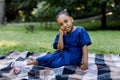 Portrait of little cute happy African girl in blue dress, sitting on checkered blanket in summer park, having a picnic Royalty Free Stock Photo