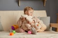 Portrait of little cute toddler blonde baby girl sitting on sofa and hugging soft toys, playing alone at home in living room, Royalty Free Stock Photo