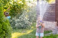 Portrait of little gardener girl, she is watering flowers on the lawn near cottage. Cute girl holding hose sprinklers Royalty Free Stock Photo