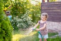Portrait of little gardener girl, she is watering flowers on the lawn near cottage. Cute girl holding hose sprinklers Royalty Free Stock Photo