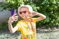 Portrait of little girl child taking a selfie by smartphone in summer park. A cheerful little girl in a yellow dress Royalty Free Stock Photo