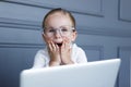 Portrait of little girl in eyeglasses behind laptop computer, making mimic with her hands, over on a grey background Royalty Free Stock Photo