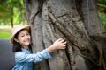 Portrait of little girl in hat with smiling,hugging large tree trunk with arms around tree and looking at camera at outdoor park, Royalty Free Stock Photo