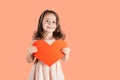 Portrait of little girl with long curly hair hold in hands big red paper heart smiling on peach background. Copy space. Royalty Free Stock Photo