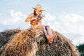 Portrait of little girl playing small guitar ukulele, plunking on haystack in field near trees. Wearing dress and hat. Royalty Free Stock Photo