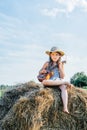 Portrait of little girl playing small guitar ukulele, sitting on haystack in field near trees. Wearing dress and hat. Royalty Free Stock Photo