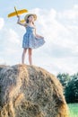 Portrait of little girl playing, standing on haystack in field. Playing flying toy airplane.Light sunny day. Low angle Royalty Free Stock Photo