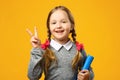 Portrait of a little girl schoolgirl on a colored yellow background. A child holds a book and shows a sign of victory.