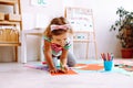 Portrait of little girl sitting near magnetic board drawing with pencil on red sheet of paper around colorful papers. Royalty Free Stock Photo