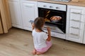 Portrait of little girl sitting near oven on floor in kitchen, open oven and looking at pie baked, kid posing backwards, wearing Royalty Free Stock Photo
