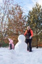 Portrait of little girl sitting on snow and mother making snowman on backyard in evening with rowan and fir trees in Royalty Free Stock Photo