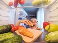 Portrait of little girl standing near open fridge full of healthy food, vegetables and fruits, and eating big croissant