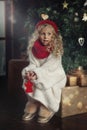 A portrait of a little girl in a white fluffy sweater and a red cap with a scarf magically sits on a box with a gift